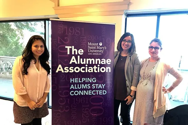 three female alumnae standing by Alumnae Association banner at event