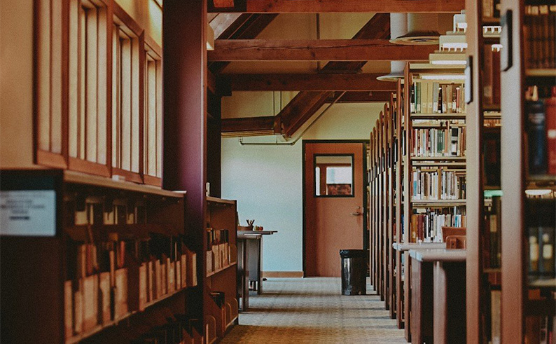 A row of library bookshelves on the right across a walkway from a wall of windows on the left.