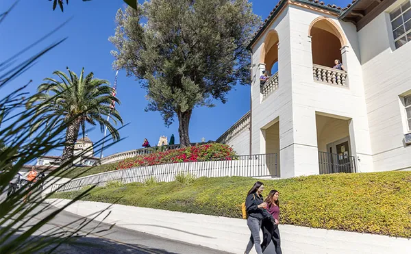 A wide shot of two students walking down a driveway with a building tower in the background