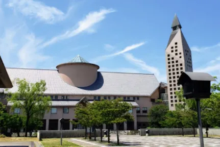 A short cream-colored building next to a tall tower with a tree-lined walkway leading to them.