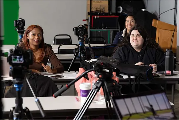 three students sitting in class listening with film equipment