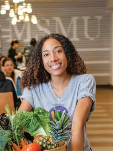 A student holding a basket of vegetables and smiling at the camera at an MSMU farmers market